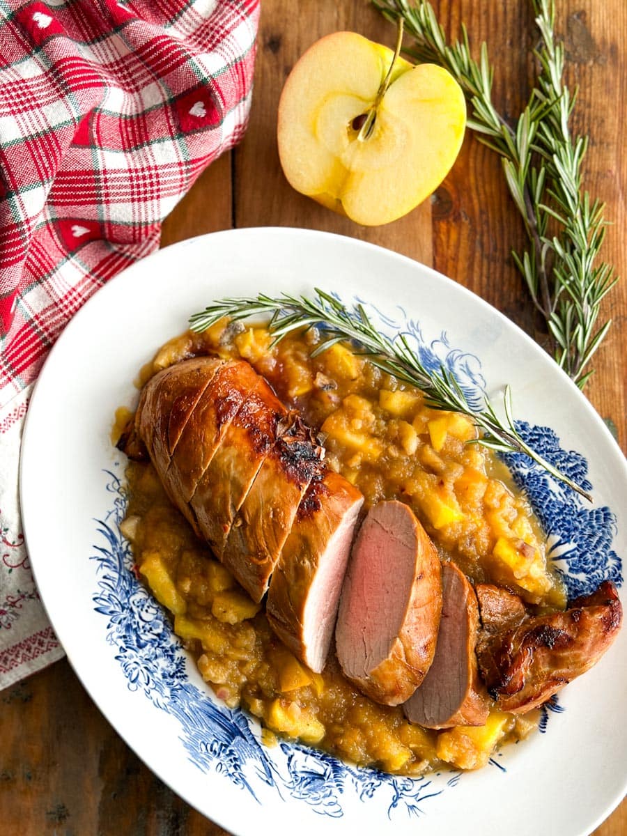 filet de porc entier grillé, sur sauce aux pommes dans une assiette de service ovale avec dessins sur table avec pomme et romarin