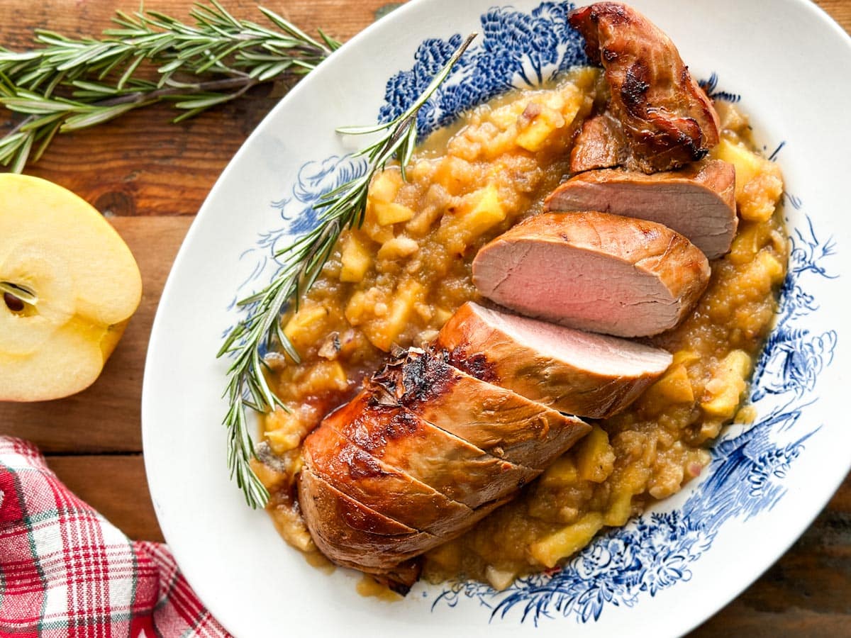 filet de porc grillé coupé en tranches sur sauce compote de pommes dans une assiette de service ovale blanche, demie pomme et romarin sur la table