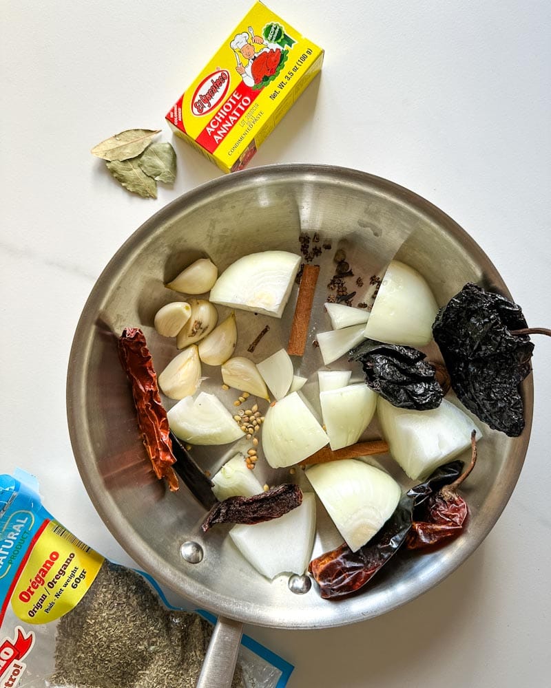 ingrédients pour birria porc effiloché: dans un poelon, oignon, ail, épices, piments, annatto et origan sur le comptoir