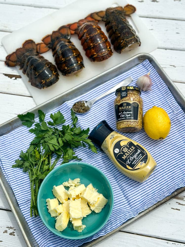 4 queues de homard, beurre, persil, moutardes Maille, citron sur une plaque sur une table en bois blanche