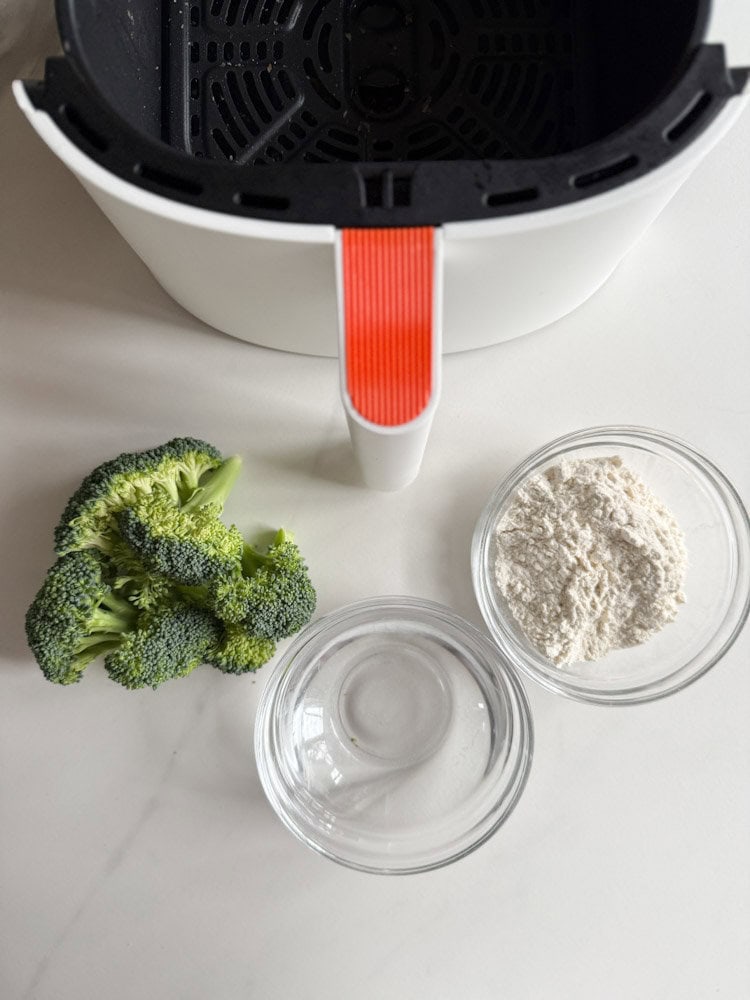 tiroir d'air fryer avec fleurons de brocoli, bol d'eau et bol de farine sur un comptoir blanc