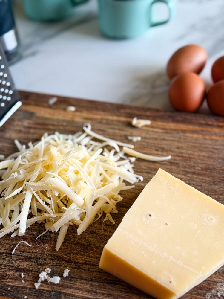 bloc de fromage comté avec fromage râpé et râte sur une planche, oeufs visibles en arrière-plan