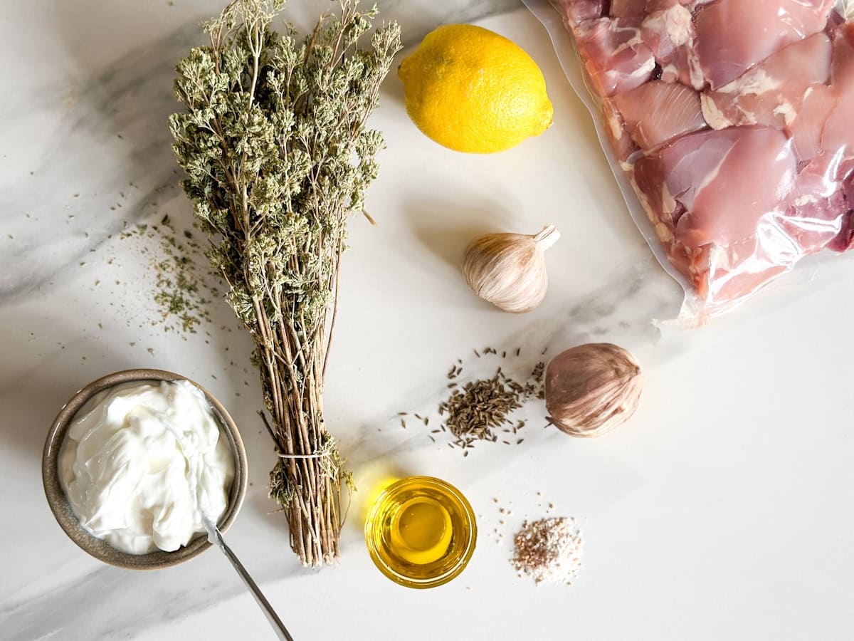 yogourt, origan en bouquet, huile, citron, ail, poulet cru et épices sur un comptoir blanc