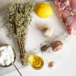 yogourt, origan en bouquet, huile, citron, gousses d'ail. épices et poulet cru dans un sac sur un comptoir blanc