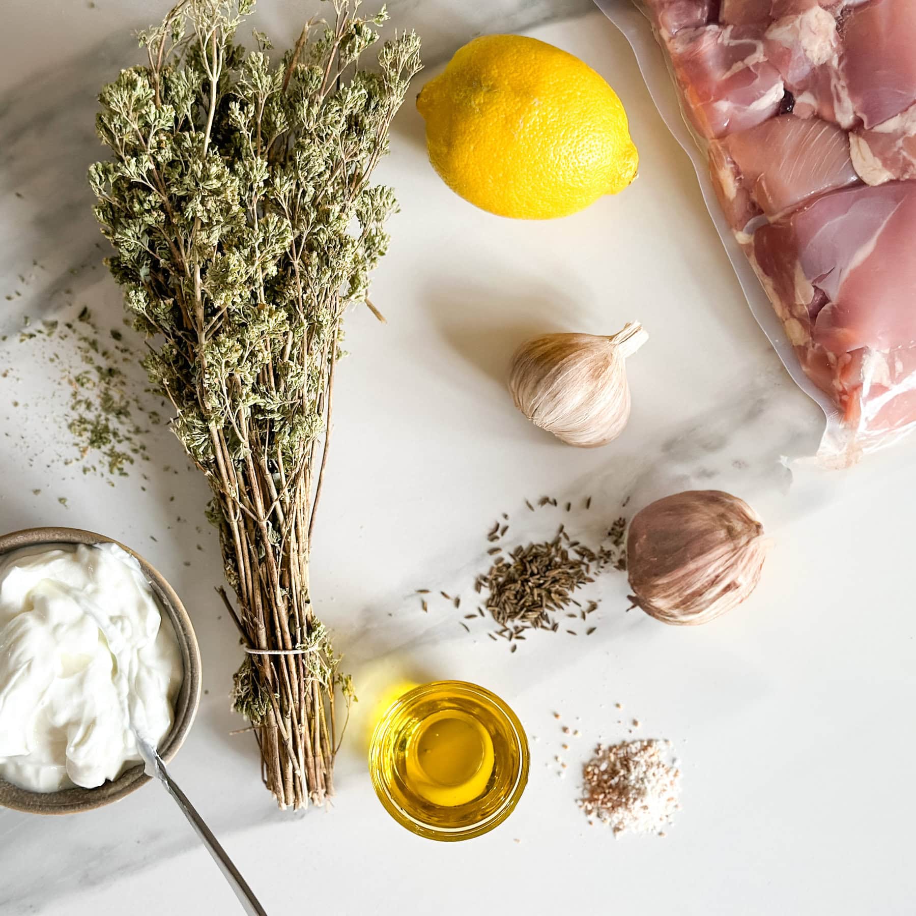 yogourt, origan en bouquet, huile, citron, gousses d'ail. épices et poulet cru dans un sac sur un comptoir blanc