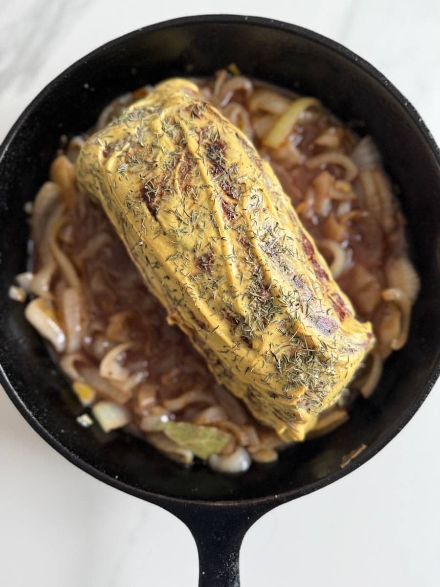 rôti français recouvert de moutarde et d'herbes sur un lit d'oignons caramélisés dans une poêle en fonte