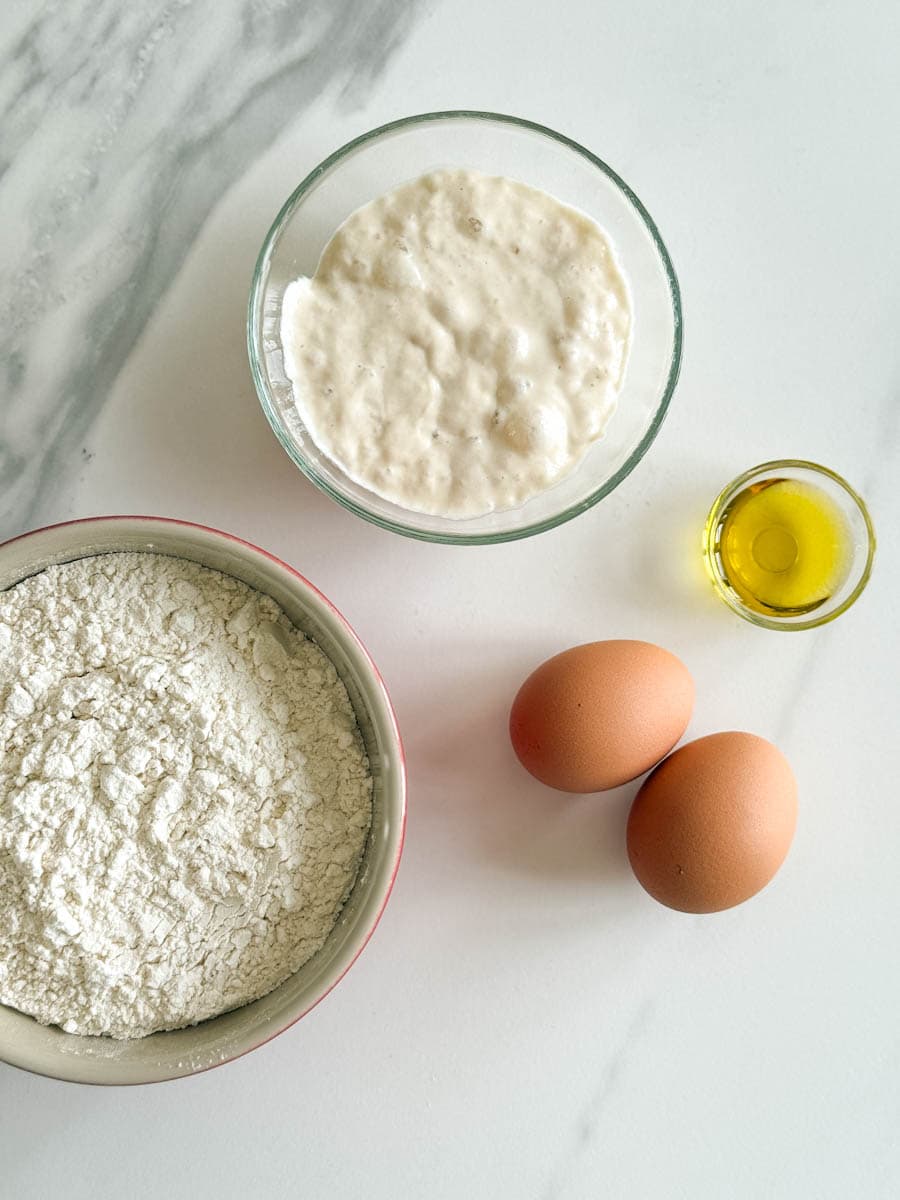 bol de farine, bol de levain, 2 oeufs et petit bol d'huile d'olive sur comptoir blanc
