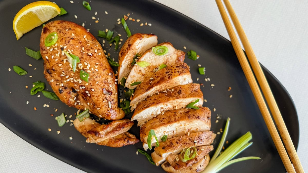 deux poitrines de poulet bien grillées dans une assiette noire avec oignons verts et graines de sésame et baguettes chinoises