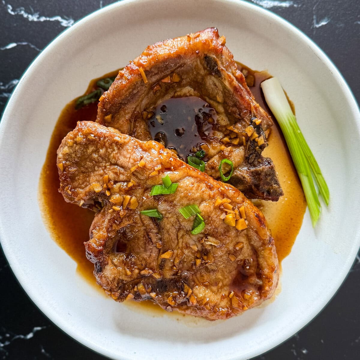 deux côtelettes de porc en sauce glacée avec oignon vert dans une assiette creuse blanche sur comptoir noir marbré