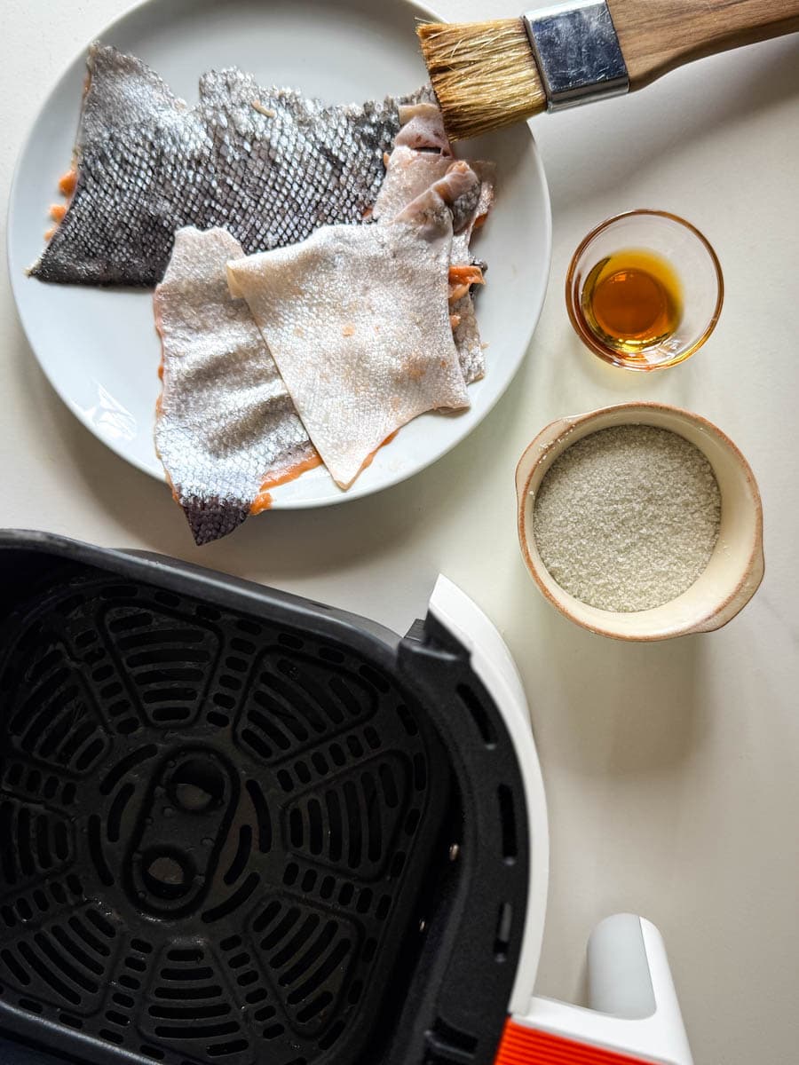 panier air fryer, peau de saumon, pinceau à badigeonner, huile de sésame et bol de sel sur comptoir blanc