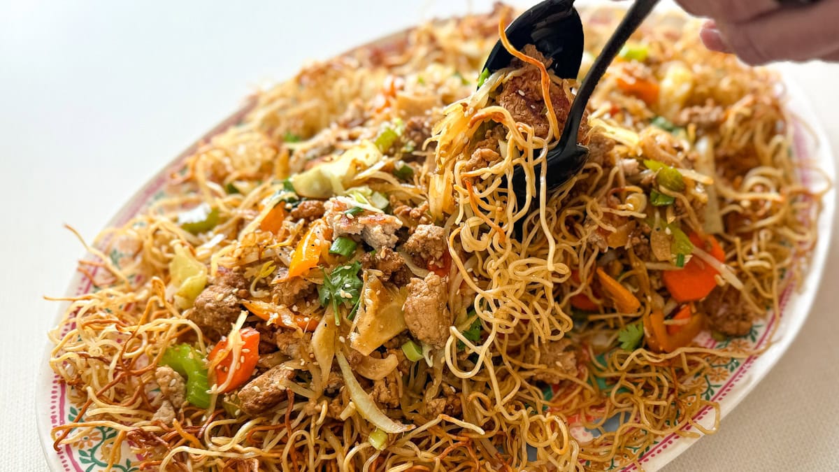 grand plat ovale de chow mein sur nouilles frites avec mains qui servent une portion avec cuillères de service