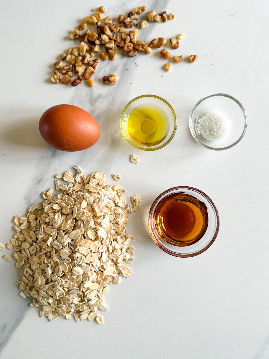 ingrédients pour granola sur comptoir blanc: noix, oeuf, sirop d'érable, huile d'olive, sel et flocons d'avoine