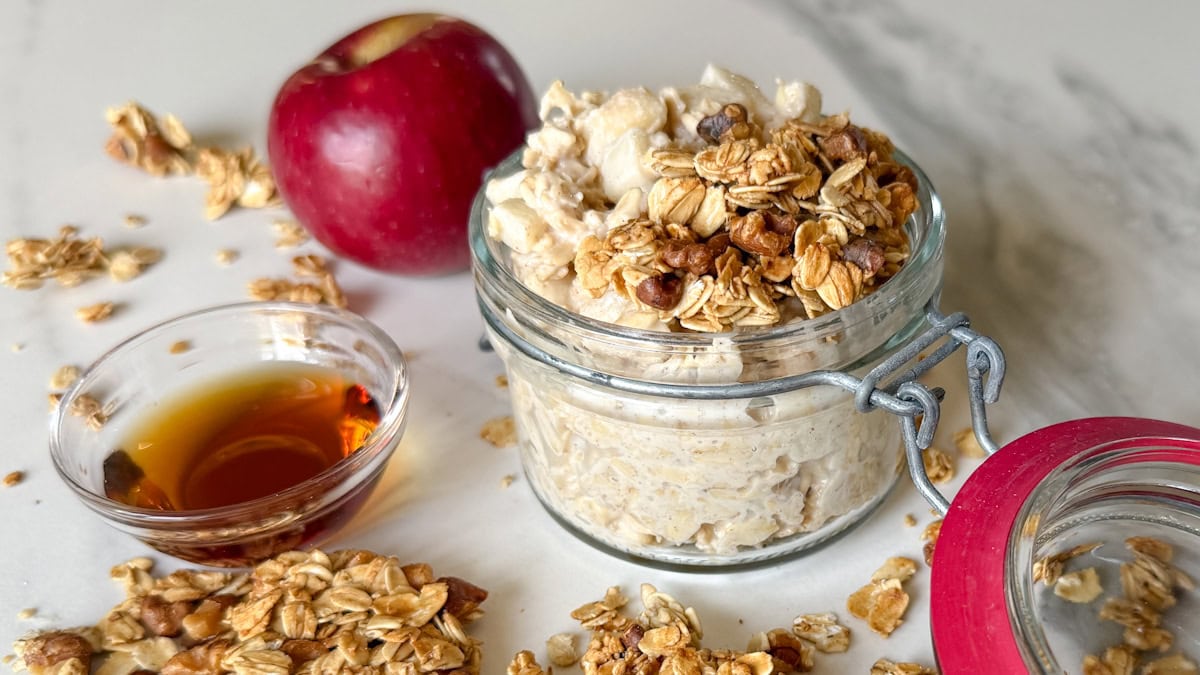 gruau oevernight dans un pot avec pomme en arrière plan, sirop d'érable et granola sur le comptoir