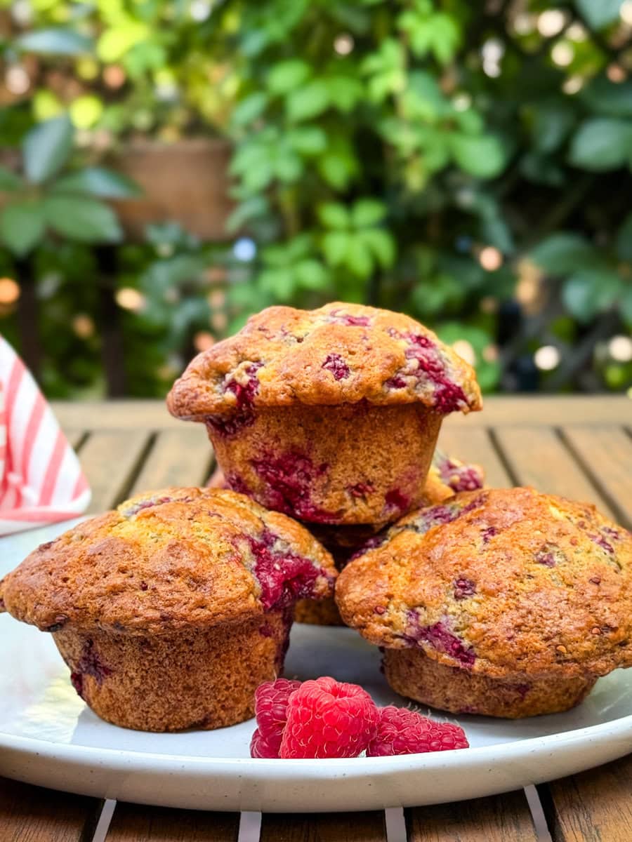 3 muffins aux framboises dans une assiette blanche avec framboise fraiche sur table en bois devant verdure naturelle