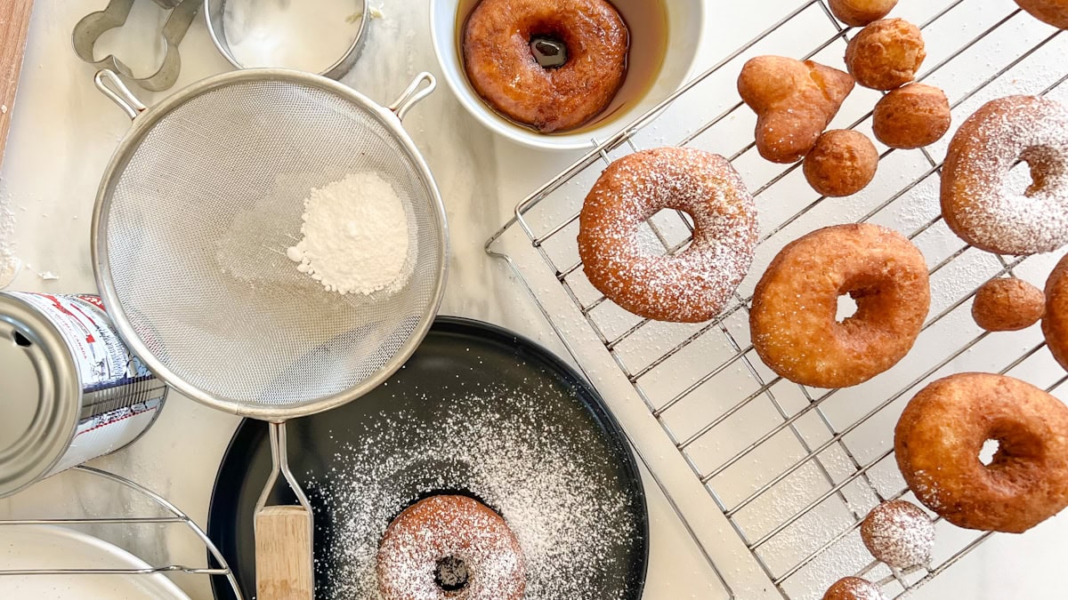 beignes aux patates sur grille de pâtisserie avec écumoir rempli de sucre à glacer, bol de sirop sur comptoir blanc marbré