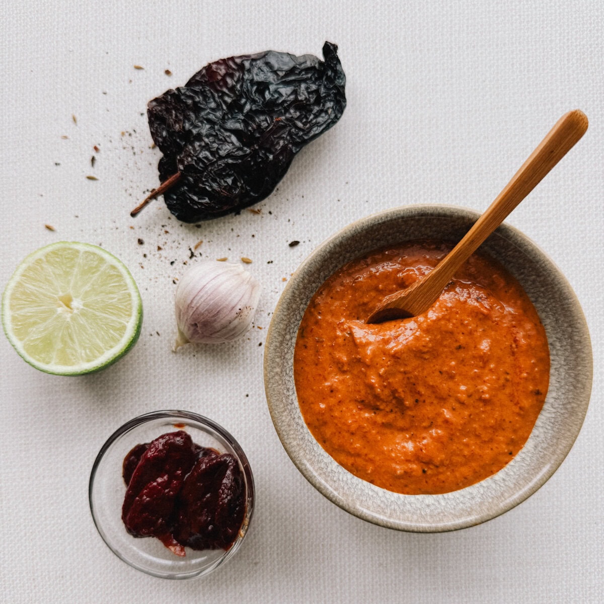 sauce et marinade chipotle dans un petit bol avec cuillère en bois, citron vert, ail et piment ancho séché à côté sur comtoir pâle