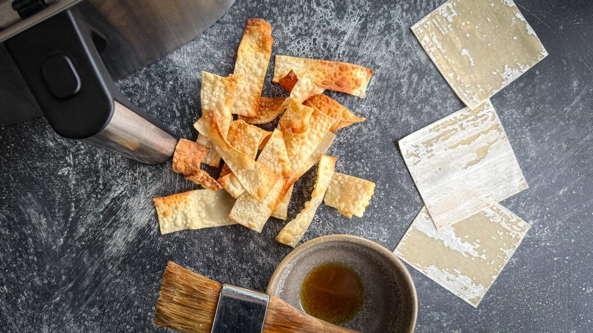 chips de wonton grillée devant tiroir du air fryer avec pâte wonton et pinceau à pâtisserie sur surface béton foncé