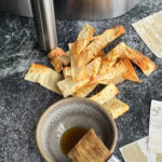 chips de wonton grillées devant panier air fryer avec petit bol d'huile grillée et pinceau à patisserie sur surface bétonnée foncée