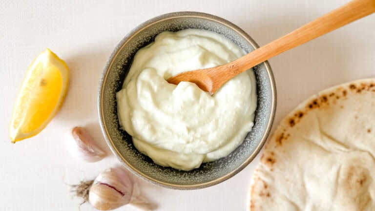 sauce à l'ail dans un petit bol avec cuillère en bois, avec gousses d'ail, citron et pain pita à côté