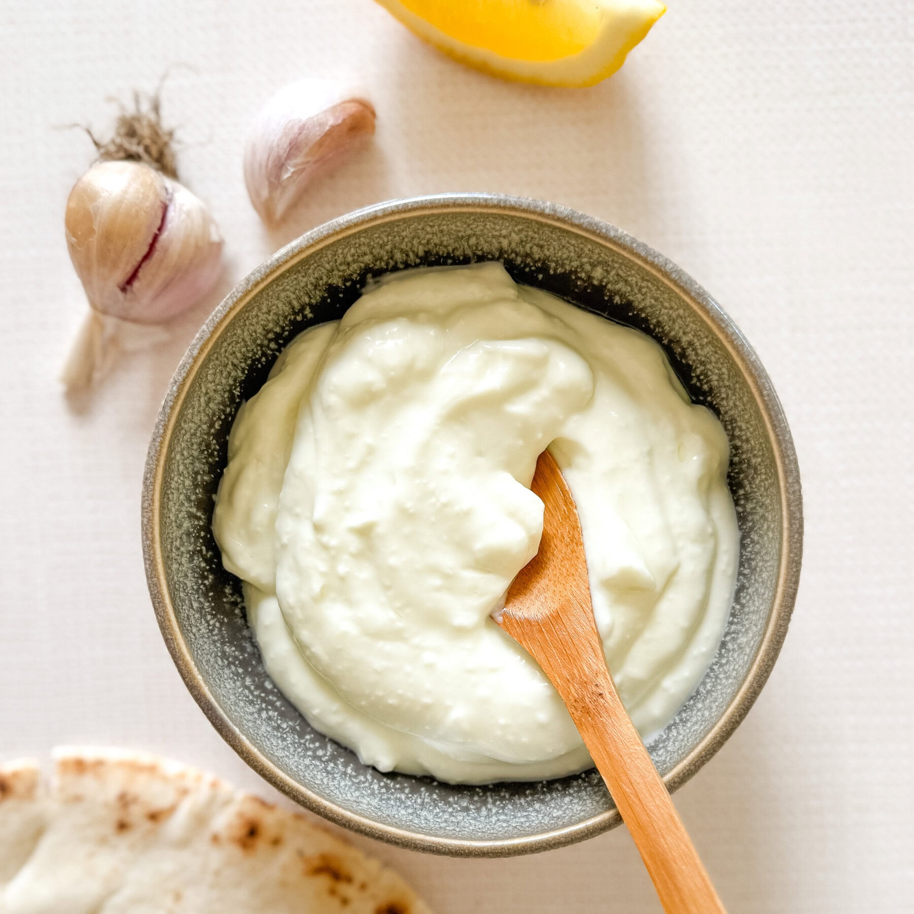 sauce à l'ail dans un petit bol en grès sur comptoir avec gousses d'ail, pain pita et quartier de citron